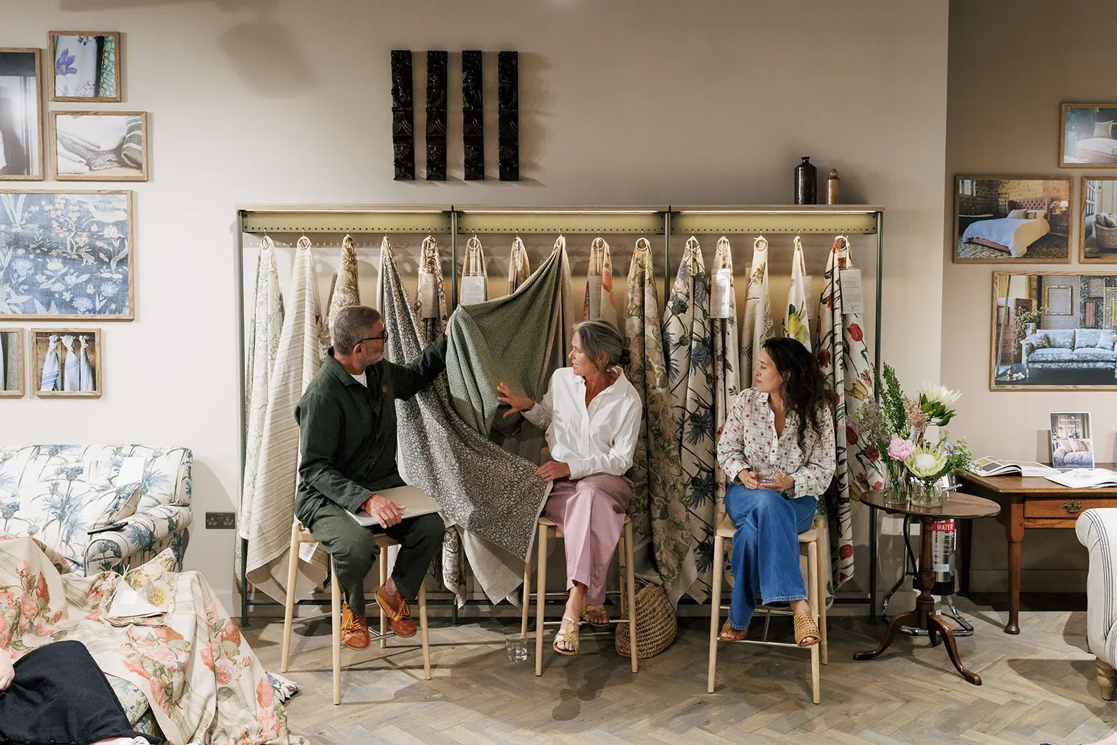 Three people sit on stools in a fabric showroom, discussing and examining patterned textiles hanging on display. The room has fabric samples, framed photos, and a cosy, creative atmosphere.